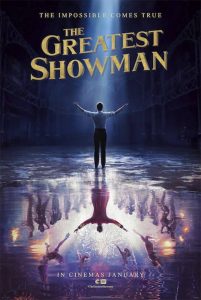 Poster for The Greatest Showman showing Barnum dressed in rolled up shirtsleeves and trousers with arms aloft to an empty room with a damp floor, reflected below it is an image of him in the same pose this time dressed in red showman's garb and surrounded by dancers and gymnasts. Tagline is "The Impossible Comes True".